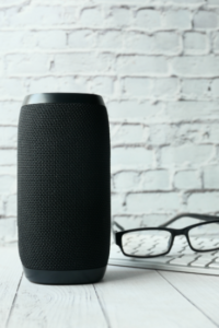 Photograph of a smart speaker against a white brick wall. Glasses and keyboard to the side.