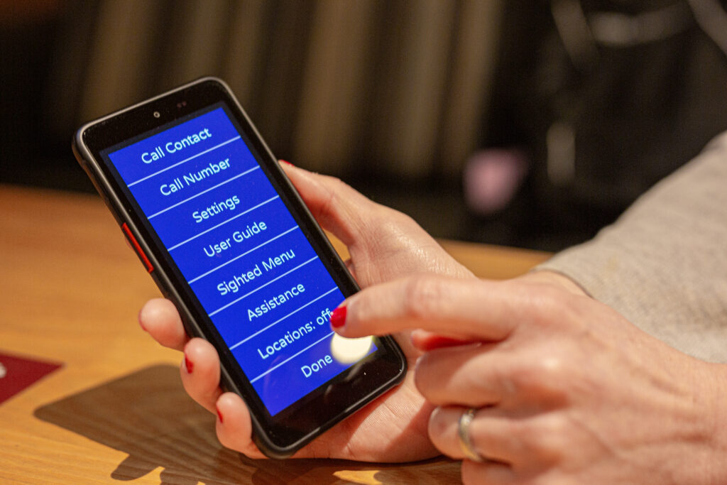 Image shows a close up of a Real Sam Pocket smart phone being held by a lady pointing at the Assistance features available on Pocket to help blind and partially sighted people