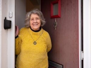 A smiling lady in her 70s wearing a bright yellow jumper opening her front door