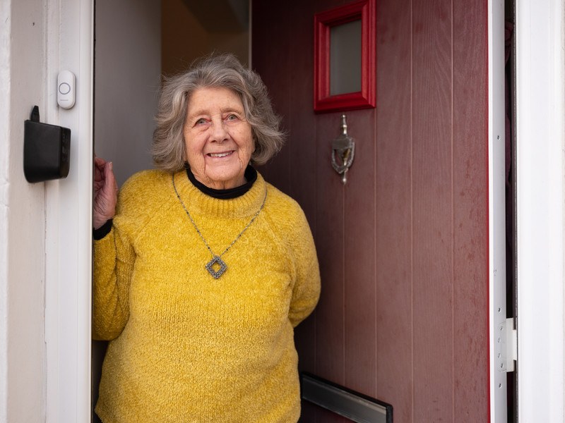 A smiling lady in her 70s wearing a bright yellow jumper opening her front door