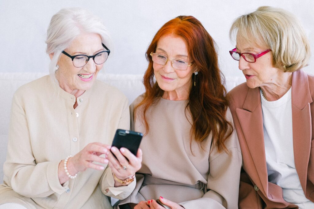 Image of three ladies sat side by side looking at a mobile phone. The ladies are each wearing glasses and smiling