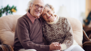 An older couple sit on their sofa smiling at the camera while holding hands