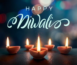 ‘Happy Diwali’ written in light blue print and fancy cursive lettering. The background has blurred lights of blues, whites, purples, and pinks. At the bottom are 5 lit Diyas, (tiny clay bowls/pots that are lit using oil and cotton string for Diwali, the festival of lights.)