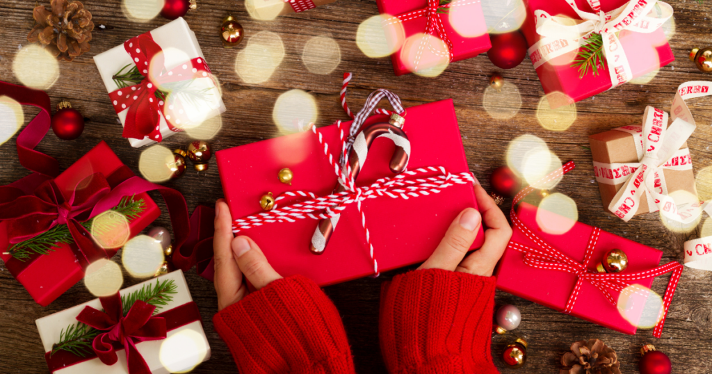 A gift wrapped in red wrapping paper and decorated with red and white, candy cane patterned ribbon, with a candy cane at the very center. The present is held, only the hands and part of the red knitted sweater sleeves are visible from the bottom of the image. The background has other finely wrapped and decorated gifts and Christmas lights. Article Image for ’12 Stocking Fillers and Gift Ideas for Blind and Visually Impaired People’