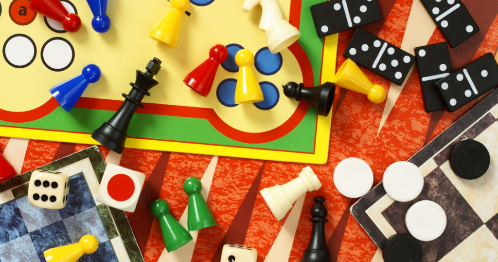 An assortment of classic games represented by their game boards and game pieces strewn about. Games include Chess, Checkers, Dominos, Trouble, etc.