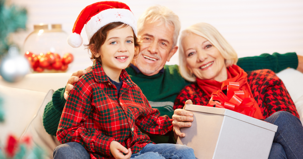 Grandparents sitting with a grandchild and holding a white package that’s about to be opened. They are all wearing holiday themed red and green outfits and the little boy is wearing a santa hat.