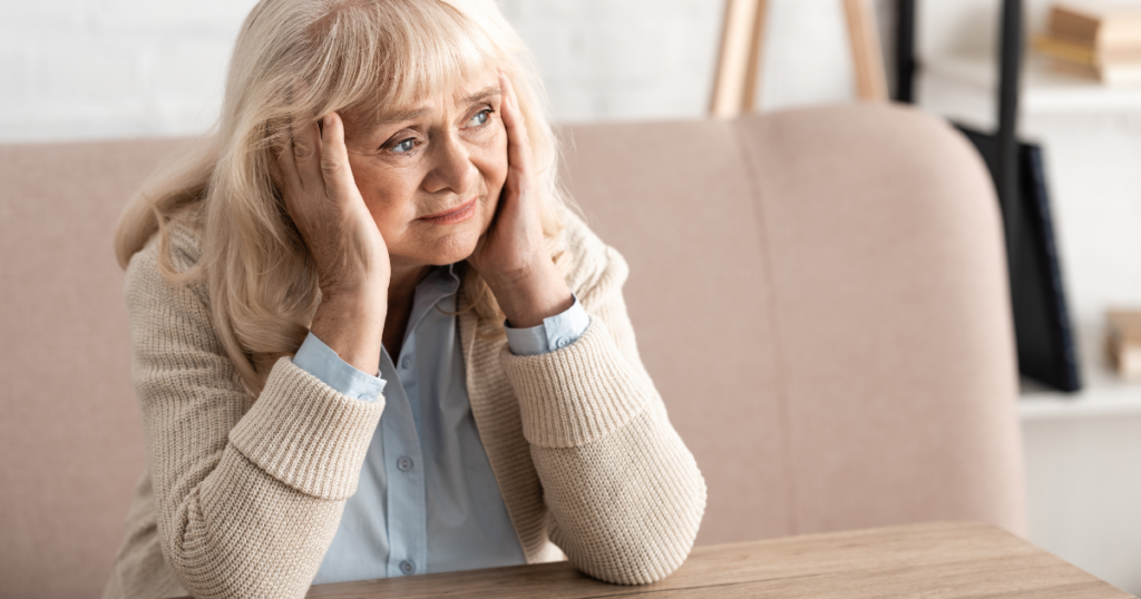 An older woman with a sad expression. She has blonde hair and greyish blue eyes looking far off. Her shoulders are hunched, her elbows are resting on a table and her face is resting on the palms of her hands.