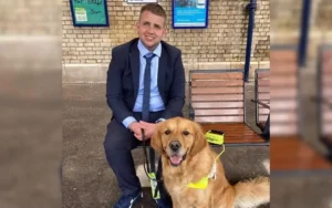 Jake is sitting on a bench at a train station with his guide dog sat at his feet. Jake is wearing a navy blue suit, with a blue shirt and tie and he's smiling at the camera