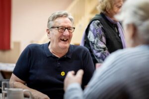 A man with white hair, galasses and wearing a navy blue polo shirt smiles at a person who is in conversation with.