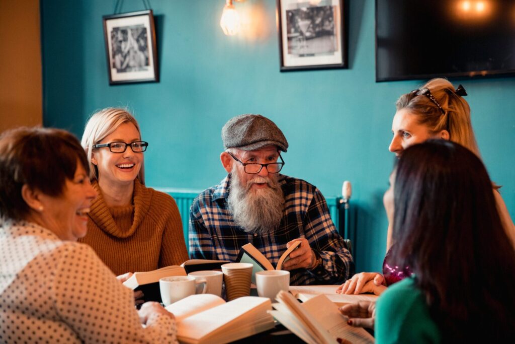 The image depicts a group of adults who appear to be engaged in a community reading circle or book club. Their physical features vary: each person has different hair colours and attire. They are seated around a table laden with books and cups. The arrangement is informal, creating a sense of community and shared activity. The individuals are positioned closely together, suggesting active interaction and conversation. The camera's angle is relatively low, framing the scene from a slightly elevated perspective, focusing on the table and the group seated around it. The foreground is filled with books and coffee mugs, while the background displays the interior of a room with teal walls and picture frames. The image demonstrates an example of a group of blind and low vision people forming connections via a book group.