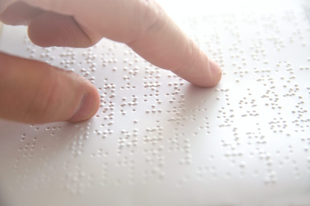 the image depicts a close up shot of a white sheet of paper with braille written on it, with a finger moving across the page. Braille is an example of accessible reading.