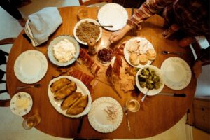 An overhead view of an autumn-style meal spread out on a round wooden table. Dishes containing mashed potatoes, gravy, sliced turkey, rolls, roasted vegetables (like broccoli), and sliced meats (possibly ham or roast beef) are arranged on numerous plates and bowls, arranged amongst fallen leaves. A hand can be seen reaching to grab a serving utensil, suggesting the food is being shared. The arrangement is somewhat informal but also organized, designed for family-style sharing. A cloth napkin is folded in the background. The lighting is diffused and warm, casting soft shadows. RealSAM's accessible cooking tips helped to make this delicious autumnal feast.