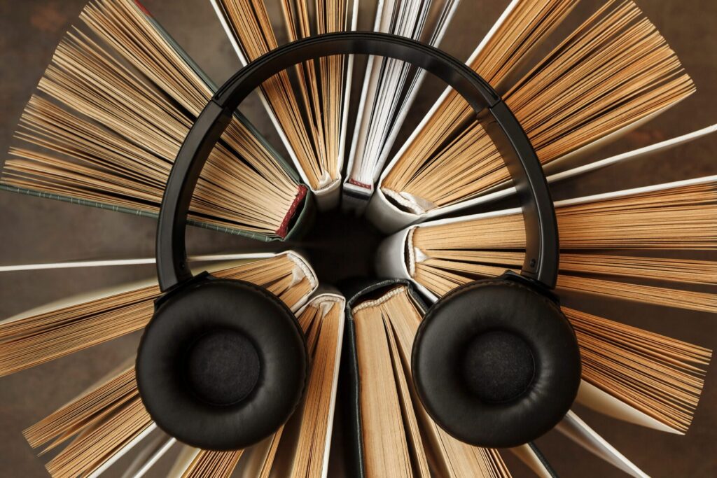The image shows a creative composition featuring a pair of black over-ear headphones resting on a circular arrangement of open books. The books are fanned out evenly, their pages splayed and facing outward to form a radial pattern, resembling a blooming flower or sunburst when viewed from above. The headphones are placed neatly at the center, their ear cups resting symmetrically on the edges of two books, symbolizing the concept of audiobooks or the fusion of literature and sound. The color palette is warm and natural, with tones of beige, brown, and cream from the book pages contrasting against the sleek black of the headphones. The background is softly blurred, keeping the viewer’s focus on the harmonious blend of reading and listening.