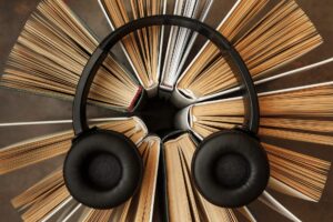 The image shows a creative composition featuring a pair of black over-ear headphones resting on a circular arrangement of open books. The books are fanned out evenly, their pages splayed and facing outward to form a radial pattern, resembling a blooming flower or sunburst when viewed from above. The headphones are placed neatly at the center, their ear cups resting symmetrically on the edges of two books, symbolizing the concept of audiobooks or the fusion of literature and sound. The color palette is warm and natural, with tones of beige, brown, and cream from the book pages contrasting against the sleek black of the headphones. The background is softly blurred, keeping the viewer’s focus on the harmonious blend of reading and listening.