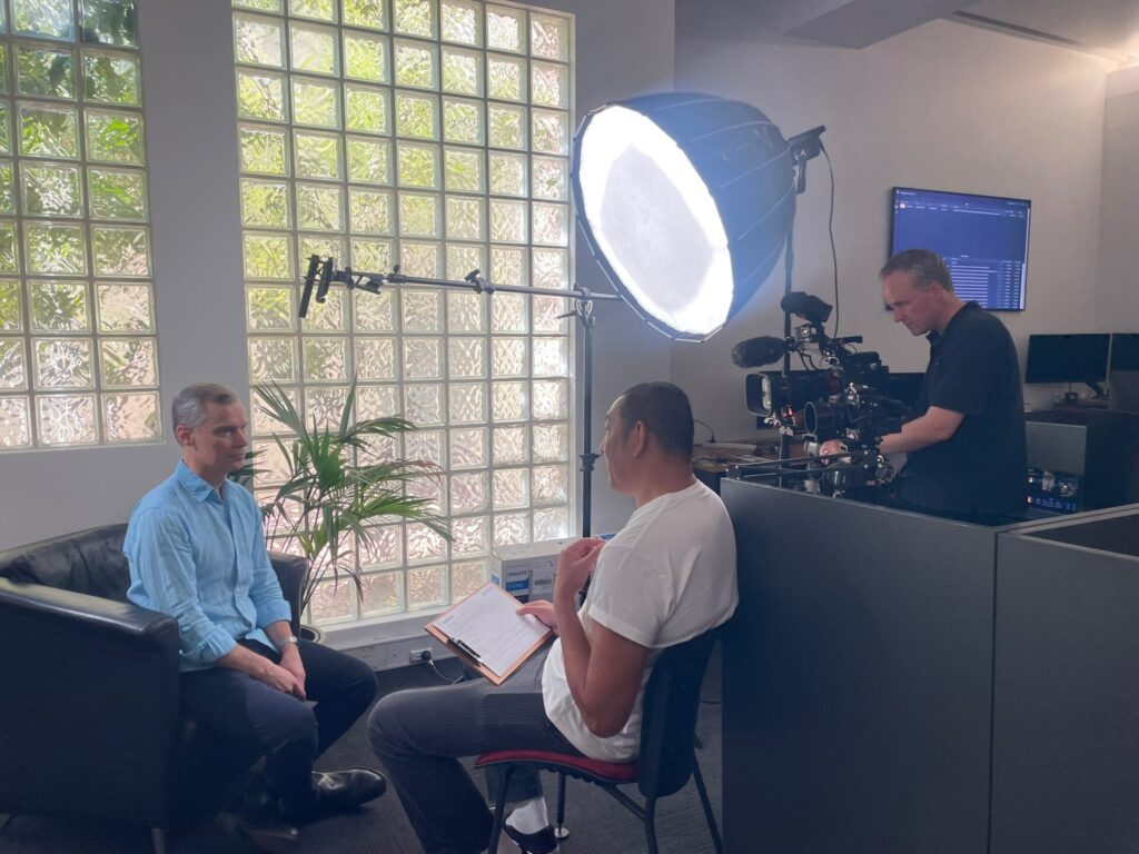 In the left of the frame the CEO of RealThing Ai, Nick, sits in a black leather chair in front of a wall of opaque glass tiles with distorted green foliage showing behind them, facing the interviewer. He is wearing a blue linen shirt. The interviewer is in the middle of the frame, sitting on a plastic chair, wearing a white t-shirt. He is in the middle of asking Nick a question. In the right-hand side of the frame, the videographer is using a large camera to capture Nick. He is surrounded by camera and lighting equipment. The overhead setup consists of a large light and a boom mic.