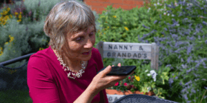 A lady with grey hair and a red top sits in her garden. She is holding a RealSAM Pocket phone and talking to it.