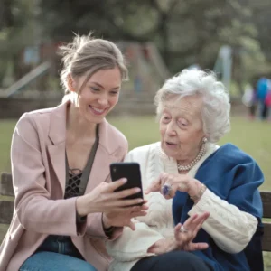 Two ladies sit on a bench and look at a mobile phone. They appear to be grnadmother and grand-daughter