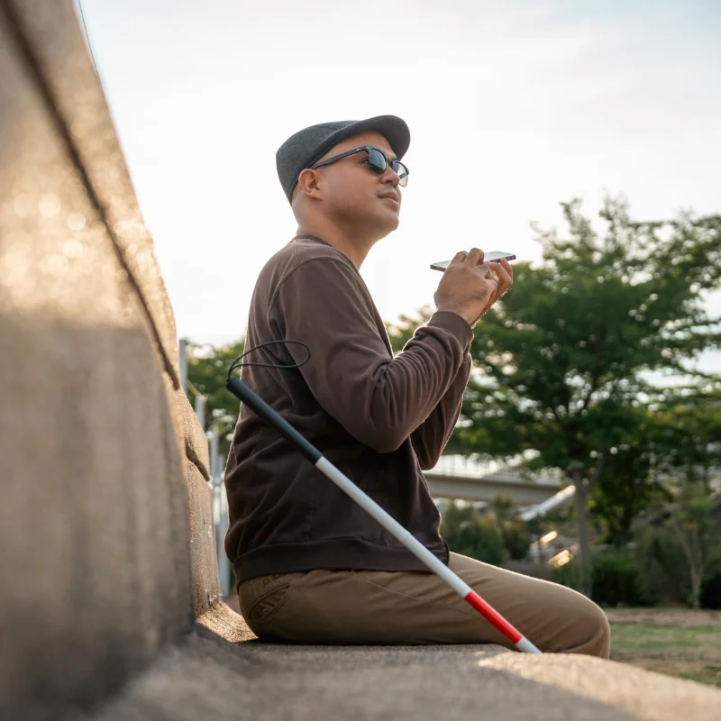 A blind man sits on a bench. He has a white can and is holding a phone