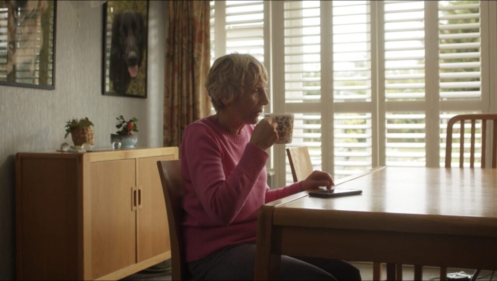 A still from the RNIB × RealSAM × Virgin Media O2 Business case-study video shows Margaret sitting at a wooden dining table in her home. She wears a bright pink jumper and holds a patterned mug close to her mouth as she takes a sip. A RealSAM device lies on the table in front of her, positioned near her right hand. Soft daylight filters through white plantation shutters behind her, illuminating the calm, homely setting. The scene captures a quiet moment of independence, reflecting how Margaret uses accessible technology in her everyday life.