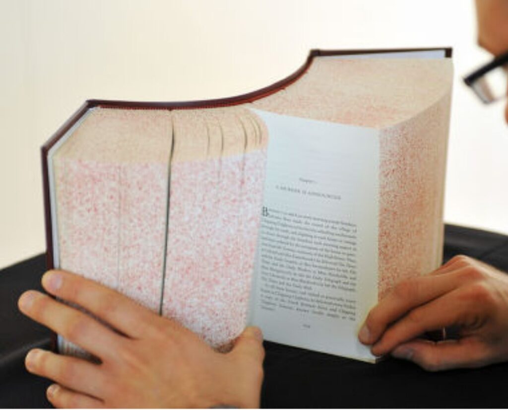 A pair of hands holds open an extremely thick book with curved, rounded edges. Most of the book’s pages are densely speckled with tiny red dots, giving the block of pages a textured, patterned appearance. Only one inner page is turned open, revealing standard printed text. The hands grip the book gently from both sides, and part of a person’s face wearing glasses is visible at the right edge of the frame. The background is softly lit and neutral, drawing attention to the unusual design of the book.