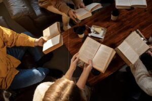 A group of people sitting around a wooden table, each holding open books. They are engaged in reading, with coffee cups beside them. The scene feels cozy and focused. The shot is from above so none of their faces are showing.