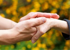 Two hands holding an elderly hand, conveying that they are helping a blind or partially sighted person. The background is blurred orange flowers, adding warmth and comfort to the scene.