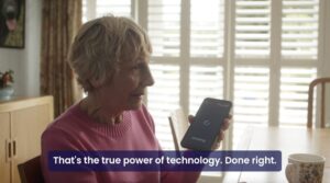 A smiling elderly woman in a pink sweater sits in a sunlit room holding a RealSAM device that displays the word "thinking." Text below reads, "That's the true power of technology. Done right" identifying RealSAM as a powerful assistive technology for vision loss.