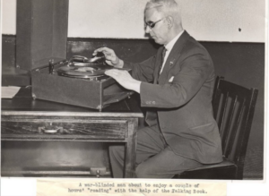 Black-and-white photo of a war-blinded man in a suit operating a talking-book phonograph, in reference to a long history of accessible reading. Below the image is written the text: "A war-blinded man about to enjoy a couple of hours' "reading" with the help of the Talking Book. Image sourced from the RNIB.